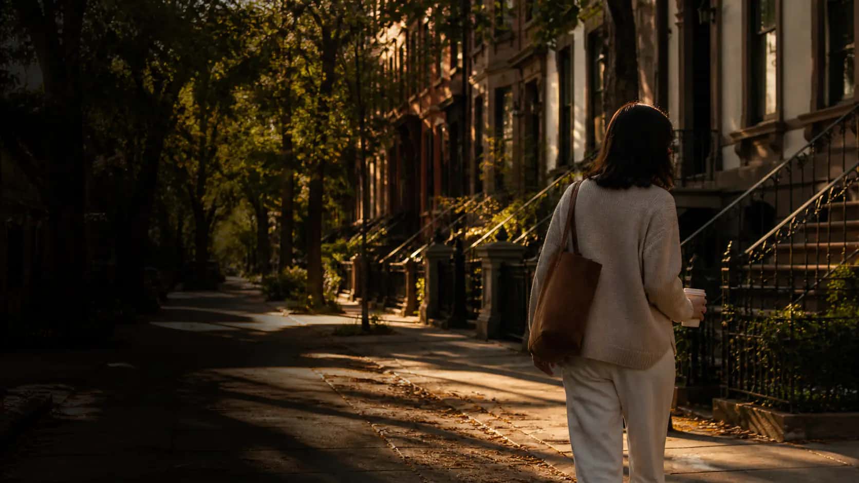 Person carrying coffee along a leafy Brooklyn street at golden hour